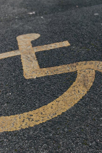 A close-up view of a yellow wheelchair accessibility symbol painted on a textured dark asphalt surface outside a residential property. Part of a house entrance is visible in the background, with a doorway, a small paved area, and some footwear seen near the entrance. A hand truck or trolley with a large black rubber wheel, used for furniture transport during home relocation, is positioned beside a doorway, supporting a stack of cardboard boxes wrapped in plastic protective film and secured with packing straps. Nearby, a blanket or moving pad covers part of furniture ready for loading into a van, which is partially visible in the background. The scene depicts the loading process at a house, involving furniture, packing materials, and moving equipment, consistent with professional removals services provided by Man with Van Blackfen, near Blackfen, as indicated on the webpage about access and parking for removals.