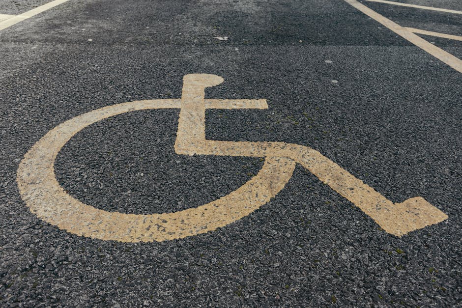 A close-up view of a yellow-painted wheelchair accessible parking symbol on a dark asphalt surface, with slightly worn and textured markings. The symbol consists of a circle representing the wheel, with an L-shape for the wheelchair frame and a smaller square for the seat area. In the background, part of a parking space with white diagonal lines is visible, indicating designated parking areas. The surface is outdoors, with natural light illuminating the scene, and the image emphasizes accessible parking spaces typically found outside residential or commercial properties involved in home relocation or furniture transport. As part of the services offered by Man with Van Blackfen for removals and moving logistics, understanding parking and access arrangements is crucial for efficient loadings and unloadings during furniture transport and packing and moving processes.