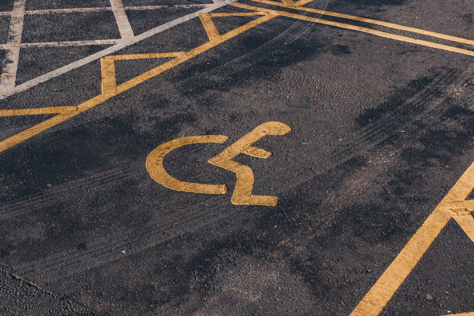 A close-up view of a yellow wheelchair accessibility symbol painted on a textured dark asphalt surface outside a residential property. Part of a house entrance is visible in the background, with a doorway, a small paved area, and some footwear seen near the entrance. A hand truck or trolley with a large black rubber wheel, used for furniture transport during home relocation, is positioned beside a doorway, supporting a stack of cardboard boxes wrapped in plastic protective film and secured with packing straps. Nearby, a blanket or moving pad covers part of furniture ready for loading into a van, which is partially visible in the background. The scene depicts the loading process at a house, involving furniture, packing materials, and moving equipment, consistent with professional removals services provided by Man with Van Blackfen, near Blackfen, as indicated on the webpage about access and parking for removals.