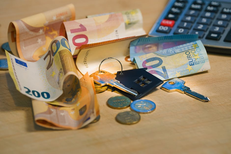 A close-up view of a scattered collection of Euro banknotes in denominations of 20, 50, and 100 euros, along with a few coins, a set of keys with a blue keyring, and a small black house-shaped keychain, all placed on a light wooden surface. Part of a calculator is visible in the upper right corner, suggesting financial or moving-related planning. The image depicts activities related to home relocation or moving logistics, where cash, keys, and financial tools are essential components. The scene is lit with warm lighting, emphasizing the details of the banknotes, coins, and moving accessories. This visual aligns with the theme of removals and the consideration of costs or hidden fees in house moving services, as highlighted in the page titled 'Are You Paying Hidden Fees in Blackfen Removals?' by Man with Van Blackfen.