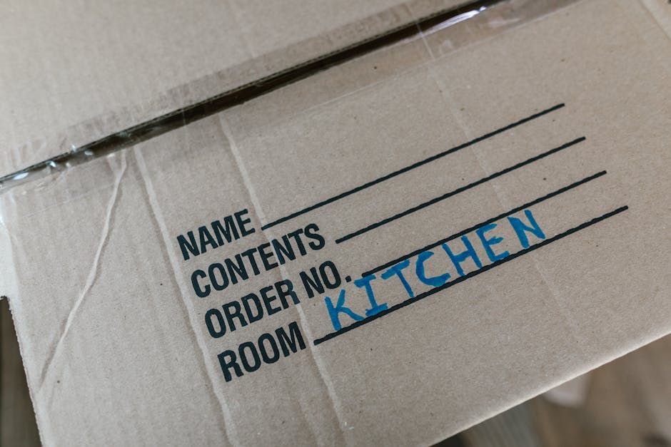 A close-up view of a cardboard box used for house removals, featuring a printed label with fields for name, contents, order number, and room designation. The label is partially filled, with the word 'KITCHEN' handwritten in blue ink in the room section. The box appears to be made of brown corrugated cardboard and is sealed with clear packing tape. It is situated inside a property, likely on a tiled or wooden floor, ready to be transported during a home relocation. Visible in the image are multiple layers of packaging materials, such as plastic wrap or film, and possibly other boxes or furniture nearby, indicative of packing and moving preparations. The scene suggests an organized loading process, where items are clearly labelled for efficient furniture transport and unpacking, consistent with the services provided by Man with Van Blackfen in house removal and clearance projects.