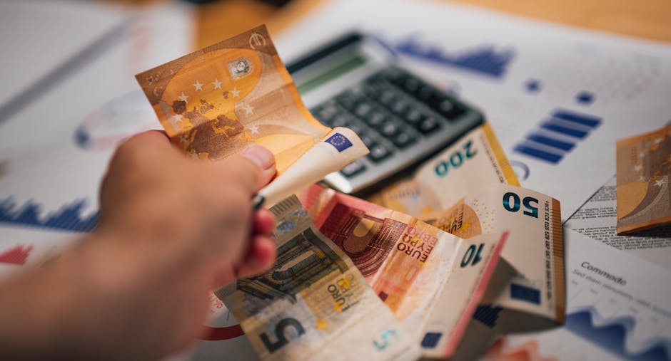 A close-up view of a scattered collection of Euro banknotes in denominations of 20, 50, and 100 euros, along with a few coins, a set of keys with a blue keyring, and a small black house-shaped keychain, all placed on a light wooden surface. Part of a calculator is visible in the upper right corner, suggesting financial or moving-related planning. The image depicts activities related to home relocation or moving logistics, where cash, keys, and financial tools are essential components. The scene is lit with warm lighting, emphasizing the details of the banknotes, coins, and moving accessories. This visual aligns with the theme of removals and the consideration of costs or hidden fees in house moving services, as highlighted in the page titled 'Are You Paying Hidden Fees in Blackfen Removals?' by Man with Van Blackfen.