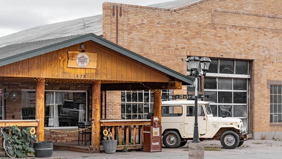 A white vintage utility vehicle is parked in front of a brick building with large window panes, on a pavement area used for a home relocation or furniture transport process. The vehicle is positioned beside a wooden porch with a gabled roof, supported by wooden beams, which features a small sign for a sandwich shop. The porch is decorated with string lights and sunflower decorations in large flowerpots, adjacent to a black metal lamppost. Cardboard boxes and wrapped furniture items are placed on the pavement near the vehicle, suggesting packing and loading activities. The background includes a red brick wall of the building, indicating an urban setting suitable for a house removals service, such as offered by Man with Van Blackfen, near Falconwood Station. The lighting is natural, with diffused daylight illuminating the scene, capturing the loading process in a typical moving environment.