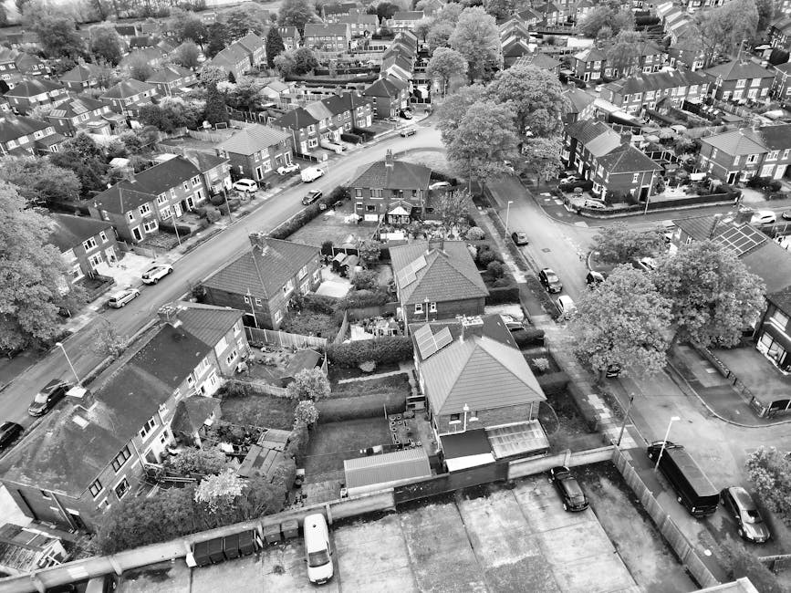 An aerial black-and-white photograph showing a residential neighbourhood with rows of semi-detached and terraced houses along curving streets, parked cars lining the roads and driveways. In the foregr