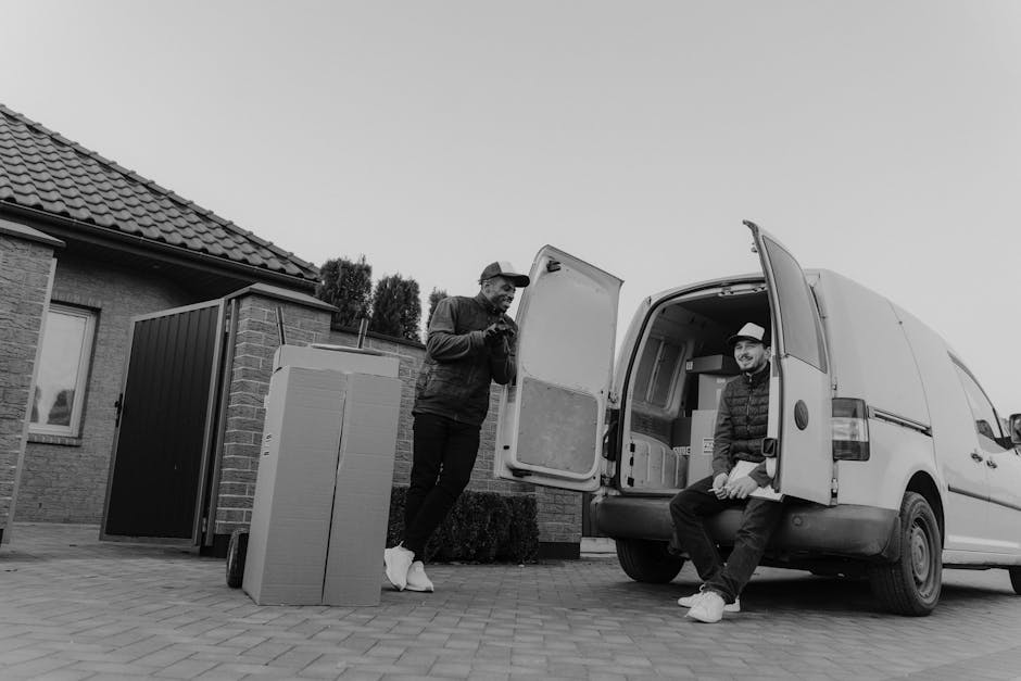 A man wearing a white polo shirt and a black cap stands inside a moving van with the rear doors open, arms crossed, and a serious expression. The van's interior is filled with stacked cardboard boxes