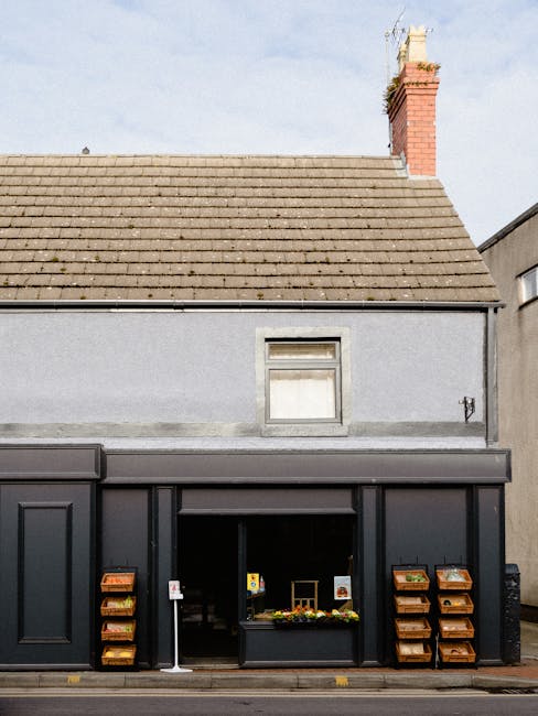 The image displays the exterior of a two-story building with a grey, textured facade. The roof is sloped and covered with brown tiles, with a red brick chimney visible at the top right corner. On the 
