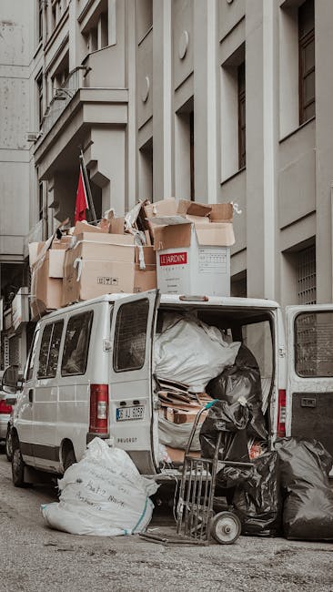 A white commercial van parked on a city street adjacent to a multi-storey building with architectural details and multiple windows. The van's rear doors are open, revealing an interior filled with large black and white trash bags, cardboard boxes, and packaging materials. Several flattened cardboard boxes and plastic-wrapped items are stacked on top of the van, partly extending onto the pavement. A metal trolley is positioned nearby, loaded with black garbage bags, indicating a collection of bulky waste likely related to home relocation or moving activities undertaken by Man with Van Blackfen. Natural daylight illuminates the scene, with the environment suggesting a residential or urban setting consistent with house removals and transport of packing materials during a move or clearance process.