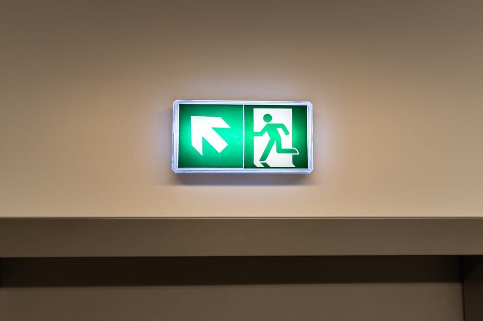 A rectangular emergency exit sign mounted on a beige interior wall, illuminated with a green background and white symbols. The sign features a white upward and rightward arrow on the left, indicating the direction to the emergency exit route, and a white figure running through a doorway on the right, representing the escape path. The sign is positioned above a lintel or door frame, with soft lighting highlighting its visibility in the indoor environment. This type of signage is commonly used during house moves or relocations to ensure safe egress routes are clearly identified for staff and residents, as managed by Man with Van Blackfen, specialists in removals and furniture transport services.