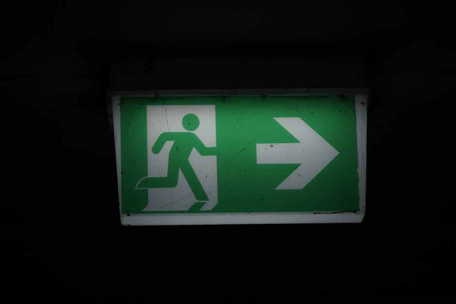 A rectangular emergency exit sign mounted on a dark wall, illuminated to display a green background with a white pictogram of a person running towards an open door, indicating the direction to an emergency escape route with a white arrow pointing to the right. The sign is intended for guiding occupants during evacuations. Although not directly related to house removals or moving services, the sign reflects safety procedures relevant to relocation environments. The setting suggests an indoor area, possibly within a residential or commercial property, where clear signage is essential for safety during home relocations or emergency evacuations. Man with Van Blackfen may include such signage as part of their comprehensive moving logistics, especially during property evacuations or when managing safety compliance during furniture transport and packing processes.