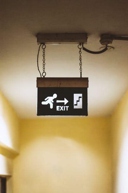 A rectangular emergency exit sign mounted on a beige interior wall, illuminated with a green background and white symbols. The sign features a white upward and rightward arrow on the left, indicating the direction to the emergency exit route, and a white figure running through a doorway on the right, representing the escape path. The sign is positioned above a lintel or door frame, with soft lighting highlighting its visibility in the indoor environment. This type of signage is commonly used during house moves or relocations to ensure safe egress routes are clearly identified for staff and residents, as managed by Man with Van Blackfen, specialists in removals and furniture transport services.