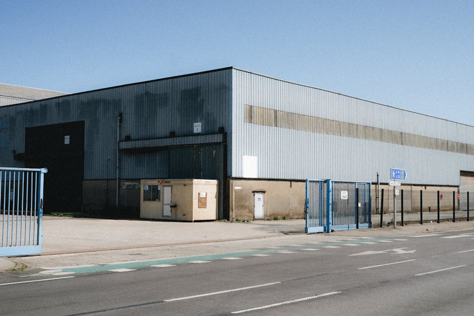 A large industrial warehouse with a corrugated metal exterior painted in shades of grey and blue, situated adjacent to a paved road with marked cycling lanes and pedestrian crossings. The building features several large black roller shutter doors, with one partially open, and small side entrances protected by metal fences and gates painted in blue. Outside, on the sidewalk, there are beige and white utility boxes and a small wooden storage box. Moving trolleys, packing blankets, and cardboard boxes may be assembled nearby in a typical home relocation context, indicative of furniture transport and packing arrangements. The exterior environment is illuminated by daylight under a clear blue sky, and the surrounding pavement is clean and well-maintained. As part of the loading process, a van belonging to Man with Van Blackfen may be positioned nearby for loading furniture and boxes into storage for a house move or property transfer. This setting reflects the logistics involved in professional removals services, including loading and transport during house relocations on Blackfen Road and DA15 properties.