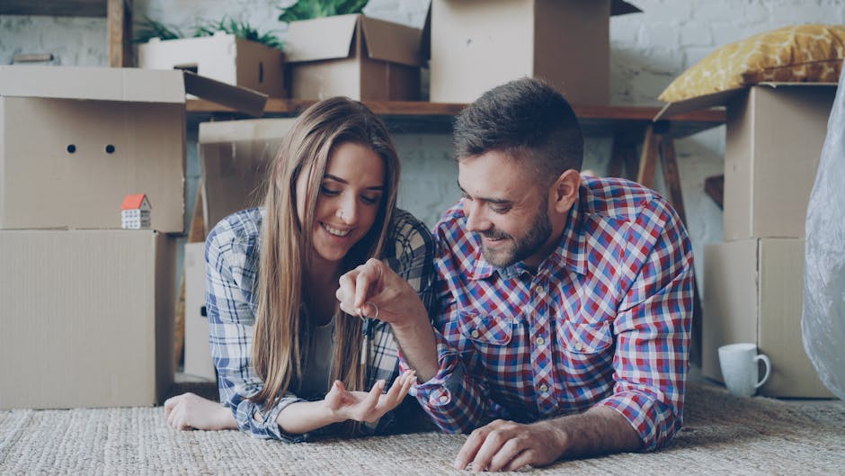 A young couple, a woman with long brown hair and a man with short dark hair and a beard, are lying on the floor of a room surrounded by moving boxes and packing materials. The woman is holding a small object in her hand, while both are smiling and looking at it. Nearby, there are cardboard boxes, some sealed with packing tape, and a miniature model house placed on one of the boxes. The background shows partially open boxes, a wooden shelving unit, a pillow, and various household items indicating a home relocation process. The scene is brightly lit with natural light, and the environment suggests they are in the midst of packing or unpacking during a house move. The presence of these packed boxes and storage items reflects the packing and moving activities associated with professional removals services like those offered by Man with Van Blackfen, supporting the theme of furniture transport and packing during house removals.