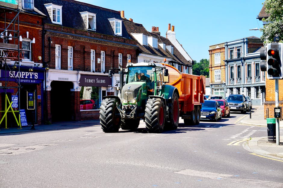 A large green tractor with an orange trailer attached is positioned at an urban street intersection during daylight hours, with the front facing the camera and the rear facing towards a row of brick a