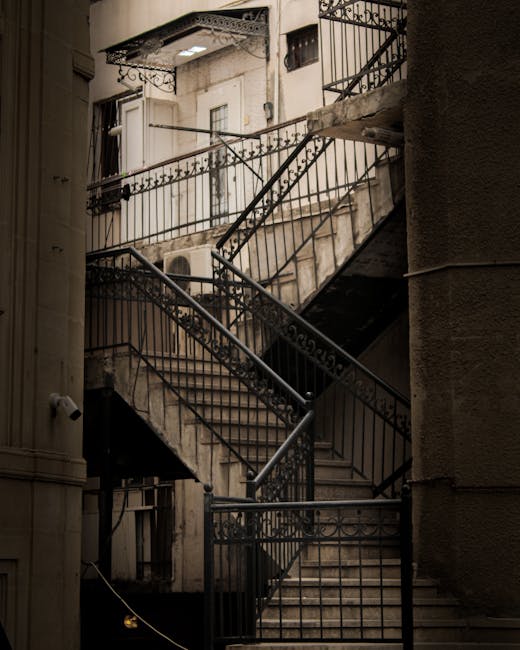 A multi-flight outdoor metal staircase with black iron railings and decorative scrollwork, attached to a brick or concrete building, leading up to a small balcony with additional railings. The staircase is situated against a light-colored, textured exterior wall, with shadows cast by the structure indicating natural light. The scene appears to be part of a residential or commercial property, with the stairs used for accessing upper floors. This type of staircase may present challenges for home relocation or furniture transport, especially in tight spaces or on narrow staircases, and professional removals services like those offered by Man with Van Blackfen are equipped to handle such environments during packing, loading, and moving processes.