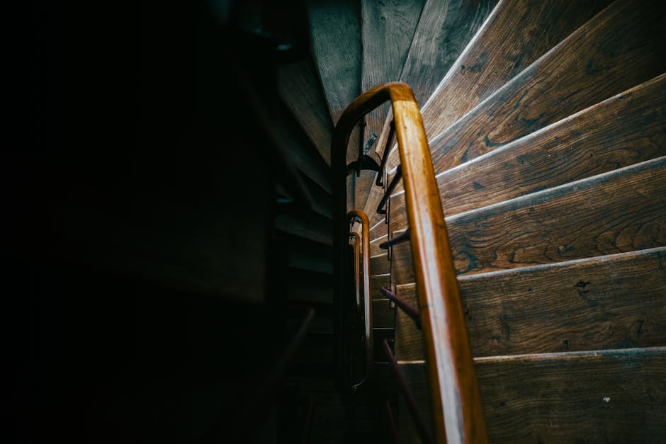 A view looking down a narrow wooden staircase inside a residential property, with dark wooden steps and a matching wooden handrail on the right side. The staircase is enclosed by dark walls, and the angle emphasizes the tight space, highlighting the steepness and narrowness of the staircase. The lighting is subdued, with natural light coming from above or an adjacent room, illuminating the wooden surfaces. This image captures the specific challenge of navigating small or confined staircases during home relocations, which [COMPANY_NAME] regularly manages as part of their professional removals services. The scene suggests a careful and organized loading process, potentially involving the transport of furniture and packed boxes up or down the staircase. The environment is typical of a residential setting needing skilled transportation solutions for furniture and belongings in limited spaces, aligning with services such as packing, moving logistics, and furniture transport, as offered by [DOMAIN].