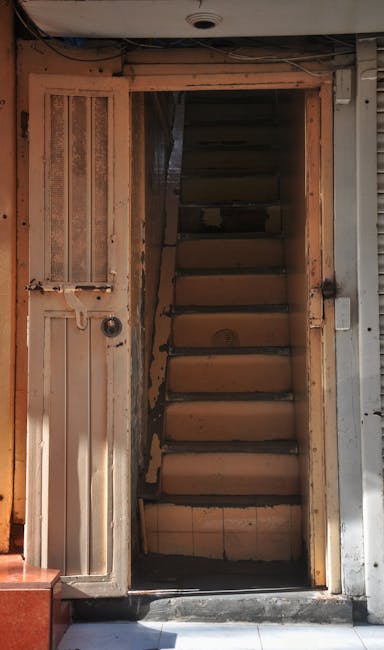A multi-flight outdoor metal staircase with black iron railings and decorative scrollwork, attached to a brick or concrete building, leading up to a small balcony with additional railings. The staircase is situated against a light-colored, textured exterior wall, with shadows cast by the structure indicating natural light. The scene appears to be part of a residential or commercial property, with the stairs used for accessing upper floors. This type of staircase may present challenges for home relocation or furniture transport, especially in tight spaces or on narrow staircases, and professional removals services like those offered by Man with Van Blackfen are equipped to handle such environments during packing, loading, and moving processes.